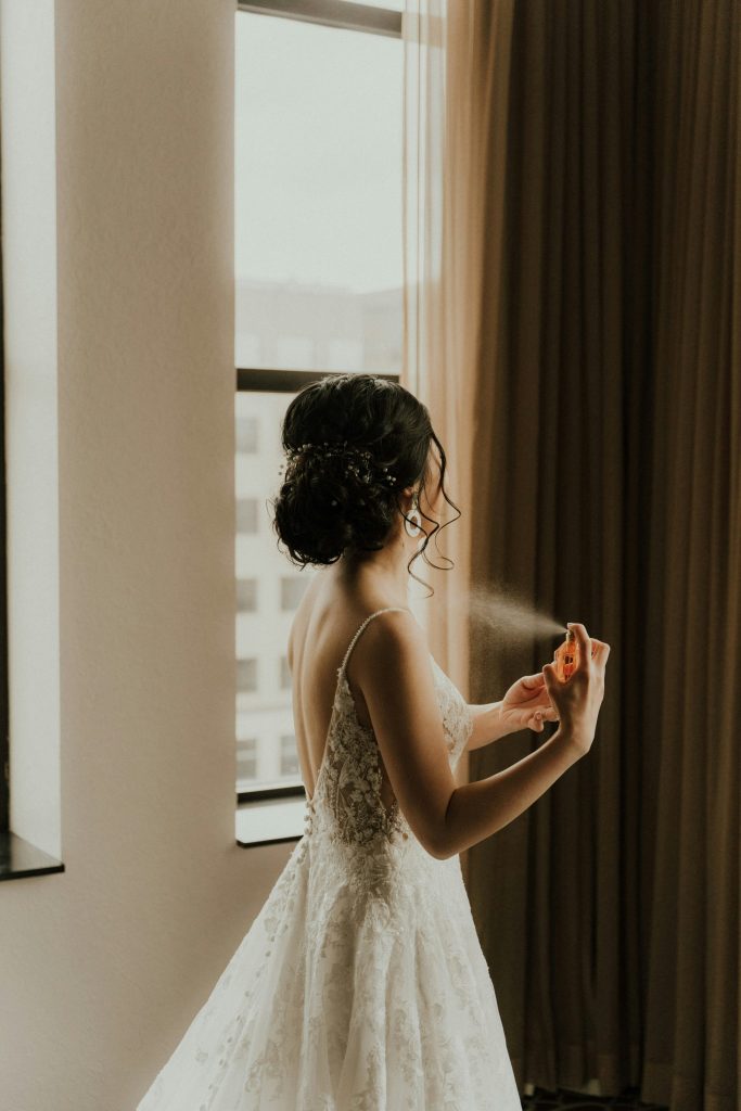 Bride in gown spraying perfume near window